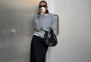 Ingrid wears a gray sweater, black woven bag, and black maxi skirt with sunglasses.