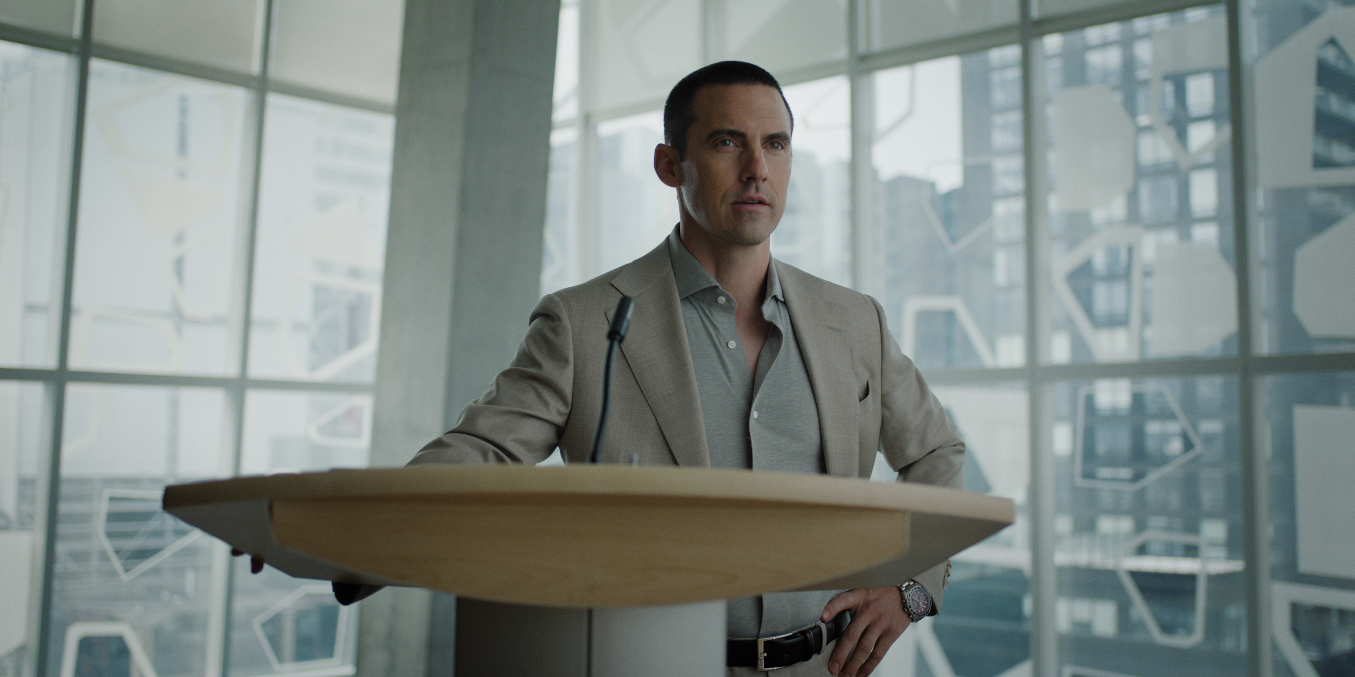 Milo Ventimiglia as hayden in I Will Find You wearing a grey suit and standing in front of a podium