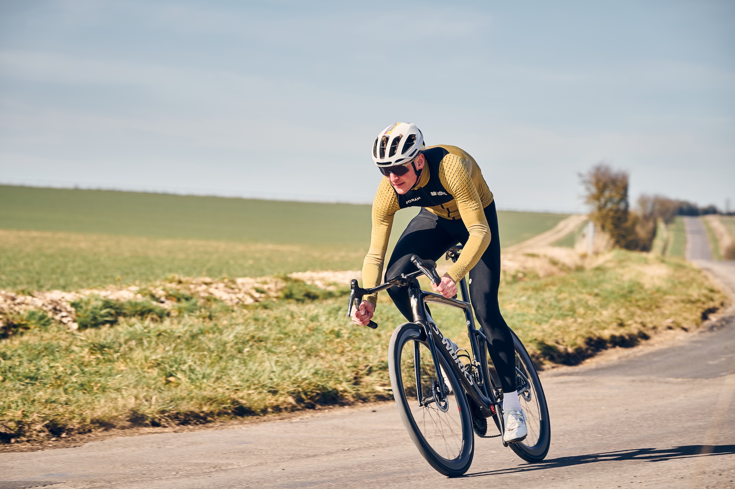 Rider in beige and black long-sleeve jersey riding hard down a slight descent, open arable land in the background