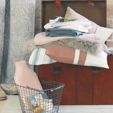 Close up of a leather trunk piled with cushions, linen and fur items. There is a basket with a hot water bottle next to the trunk.