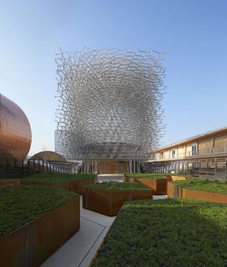 View of the UK Pavilion featuring a tall, aluminium mesh ’beehive’ structure and several patinated metal, geometric planters with low greenery under a clear, blue sky
