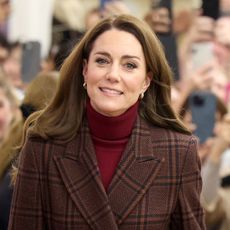 Kate Middleton has long brown hair and smiles while wearing a tartan coat and red sweater