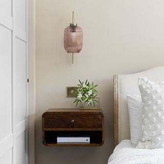 Glass and brass ribbed wall light in amethyst above dark wood, floating bedside unit with drawer and fresh flowers.