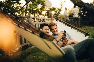 Two men sit in a hammock together and look at a phone