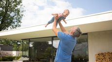 A dad lifts his toddler up into the air on the patio of his mid-century modern home.