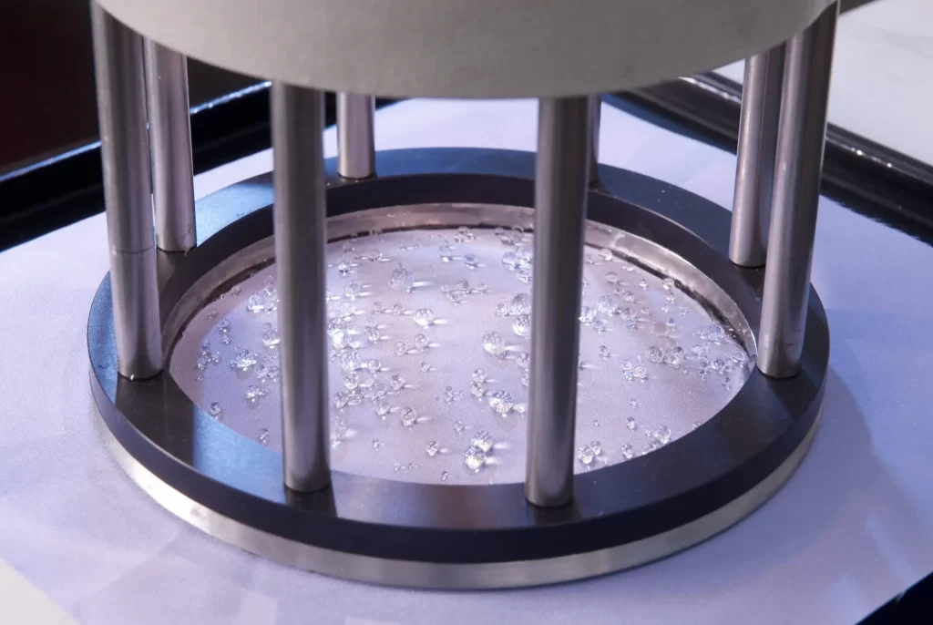 A ring supported by pillars clamps a fabric swatch on which beads of water have formed