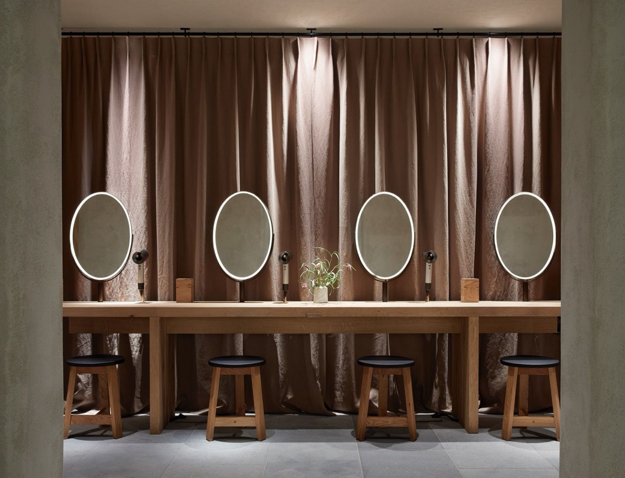 A row of wooden stools and mirrors with glowing lights in front of heavy curtains, creating a calm dressing space. Hairdryers and small plants sit neatly on the counter.