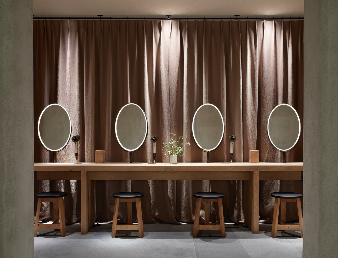 A row of wooden stools and mirrors with glowing lights in front of heavy curtains, creating a calm dressing space. Hairdryers and small plants sit neatly on the counter.