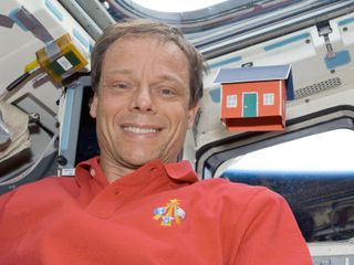 An astronaut poses with a small red and white paper house as both float in front of a window aboard the space shuttle