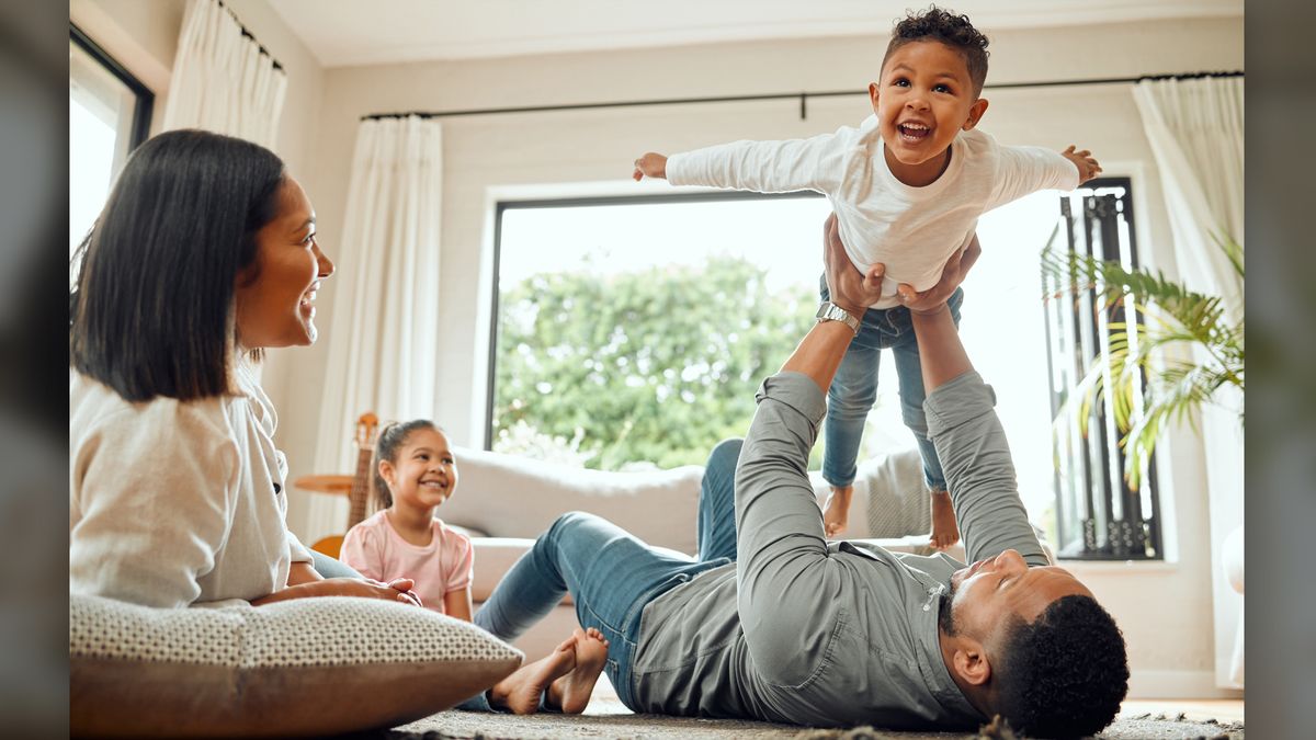 We see a mom, dad, daughter and son playing in a living room. The dad is playing &quot;airplane&quot; with his son, whose arms are outstretched. 