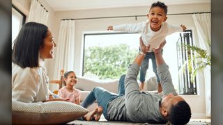 We see a mom, dad, daughter and son playing in a living room. The dad is playing "airplane" with his son, whose arms are outstretched. 