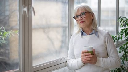 An older woman stands at the window with a cup of coffee and appears to be thinking.