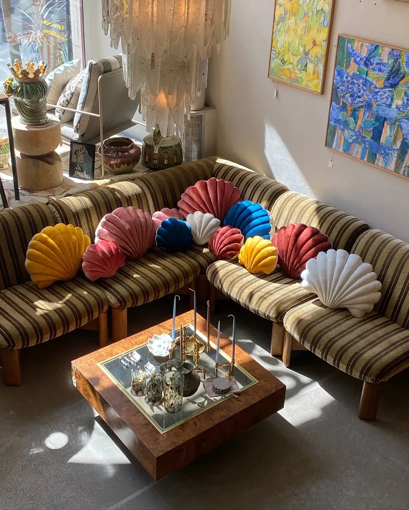 The colorful, quirky interiors of a second-hand furniture shop features coffee-brown velvety seating, shell-shaped cushions in vibrant hues, a central, square wooden coffee tables, candles, and hung-up artworks.