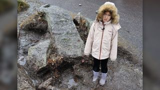 A young girl in light pinkish parka with the furry hood on her head stands near some large rocks by a concrete road where she found the dagger.