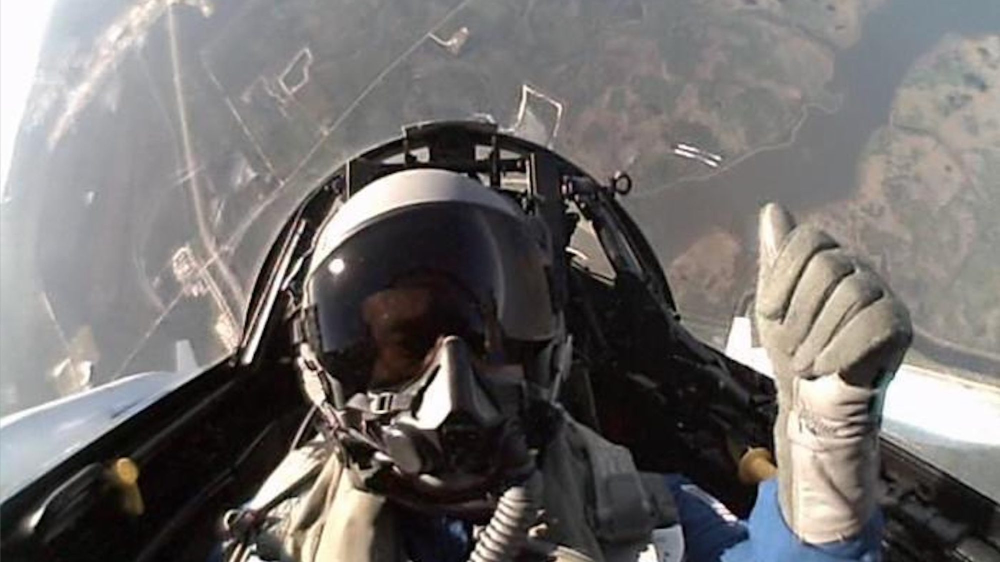 a person in a helmet gives a thumbs-up from inside a jet cockpit, with the ground far away in the background.