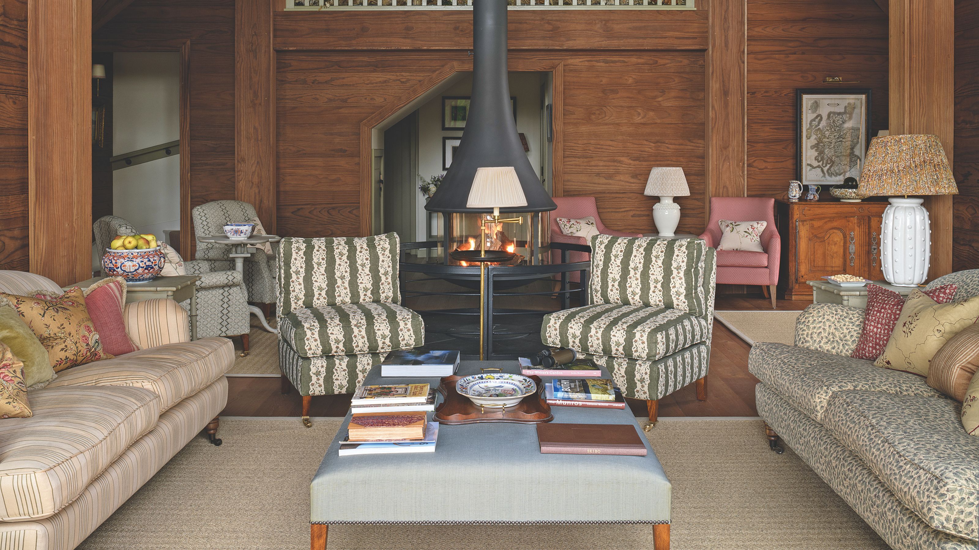 A cozy wood-paneled living room featuring a modern black hanging fireplace, striped armchairs, a blue tufted ottoman, and mismatched floral textiles.