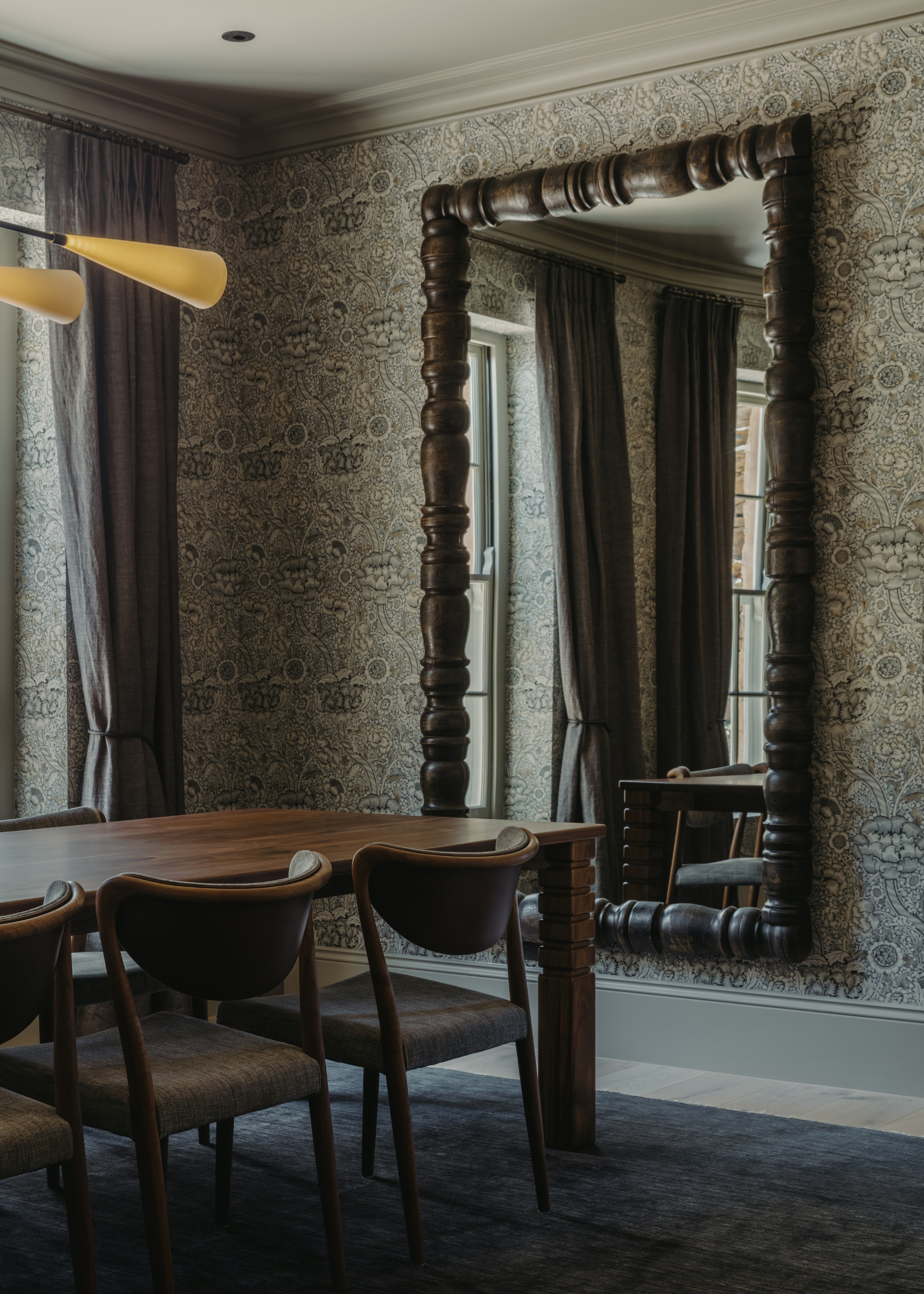 A dining room with patterned wallpaper, a textured wooden frame mirror, a wood dining set, a cozy area rug, and overhead lighting