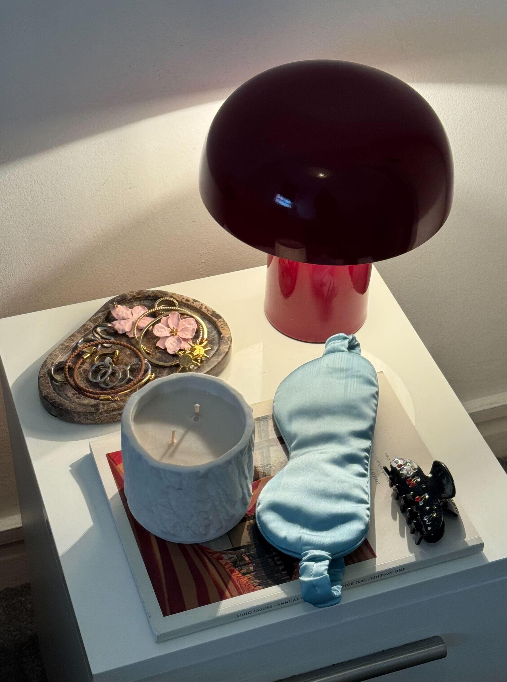 A white square nightstand with a dark red oxblood table lamp, a marble jewelry tray, a white candle, and a light blue eye mask on it.