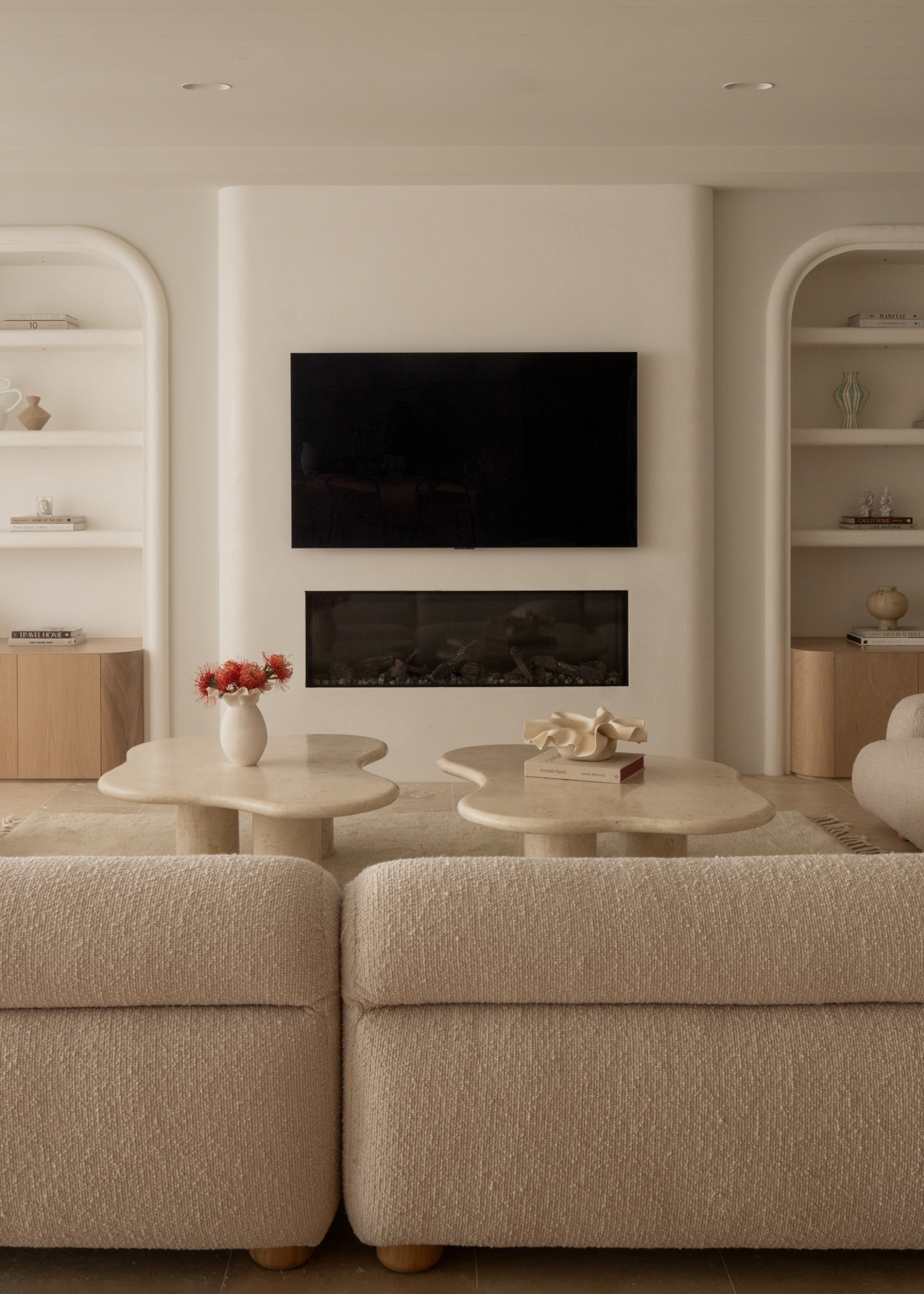 A living room with a mounted tv above a fireplace in between symmetrical arched open shelves and in front of a pair of curved marble coffee tables with vases by a beige textured couch