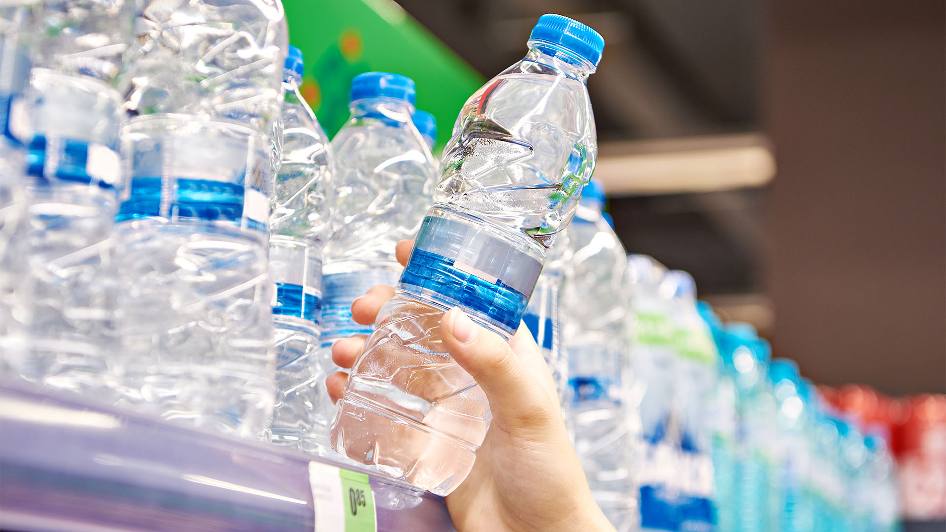 A hand reaches up to pick a plastic bottle of water from a shelf full of bottled water in a supermarket