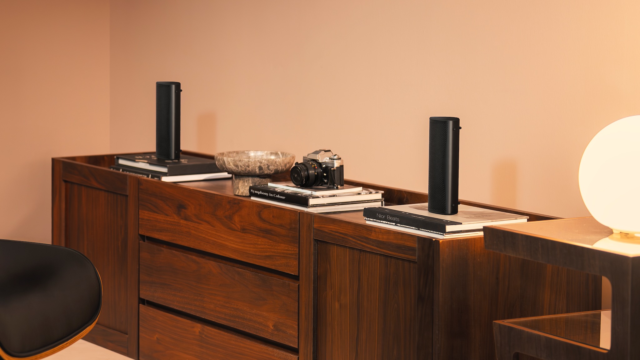 Image of two Muo speakers atop a wooden desk with a camera between them and books underneath them