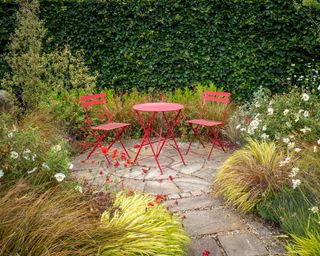 Secluded area of garden with small patio and two red chairs with metal table surrounded by summer flowers and grasses