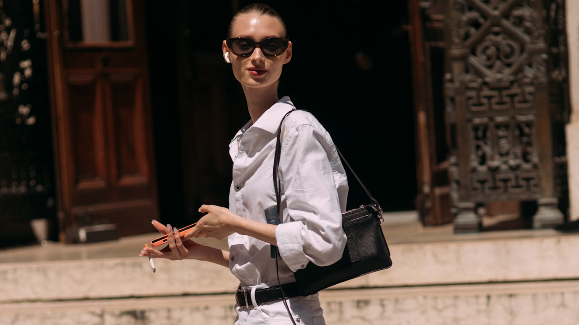 Fashion week attendee wearing sunglasses, white button down shirt, shoulder bag, and white shorts