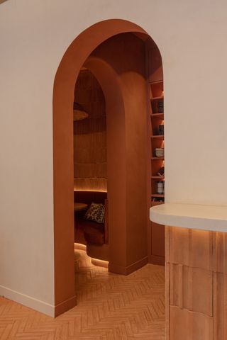 A modernly furnished restaurant in the tones of terracotta and pale pink features an arched, doorless entrance to a private dining room with suffused lighting, brick walls, and bespoke banquettes.