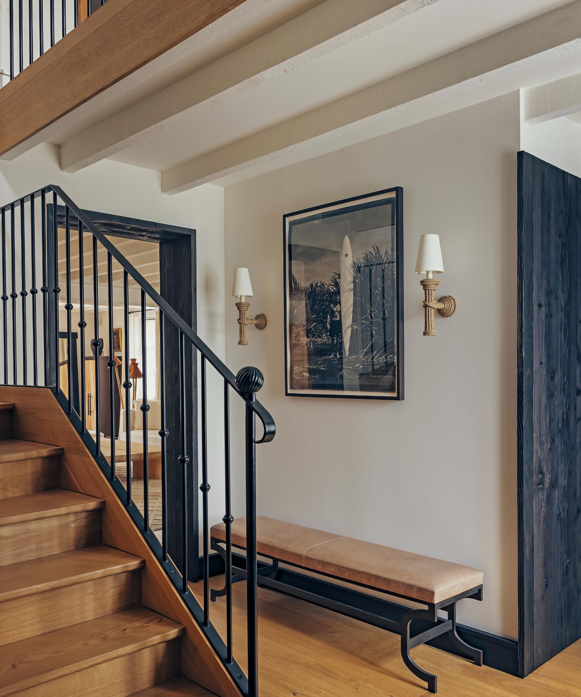the entryway to a rustic beachside home in Biarritz with wooden beams, oak staircsae with black metal railings and a small bench with surf photographer hung above