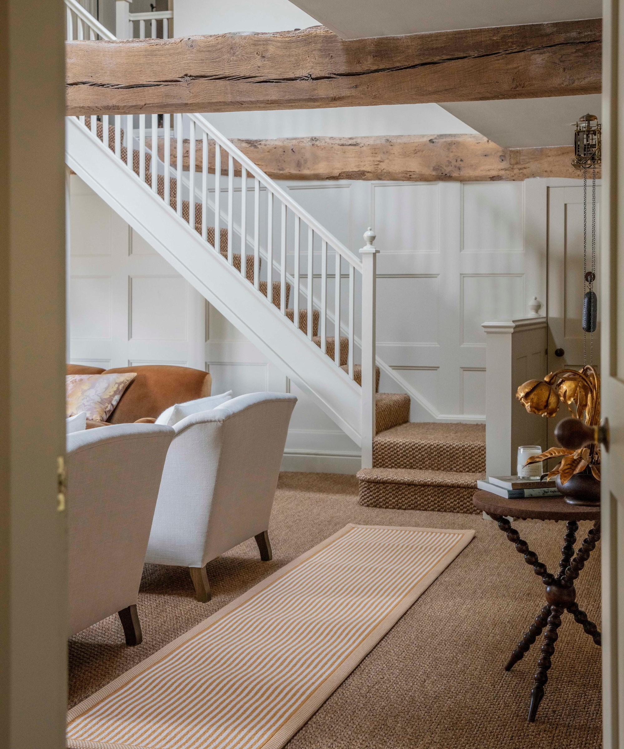 Staircase with white balustrade and exposed wood beam, striped runner over sisal carpet, armchairs below.