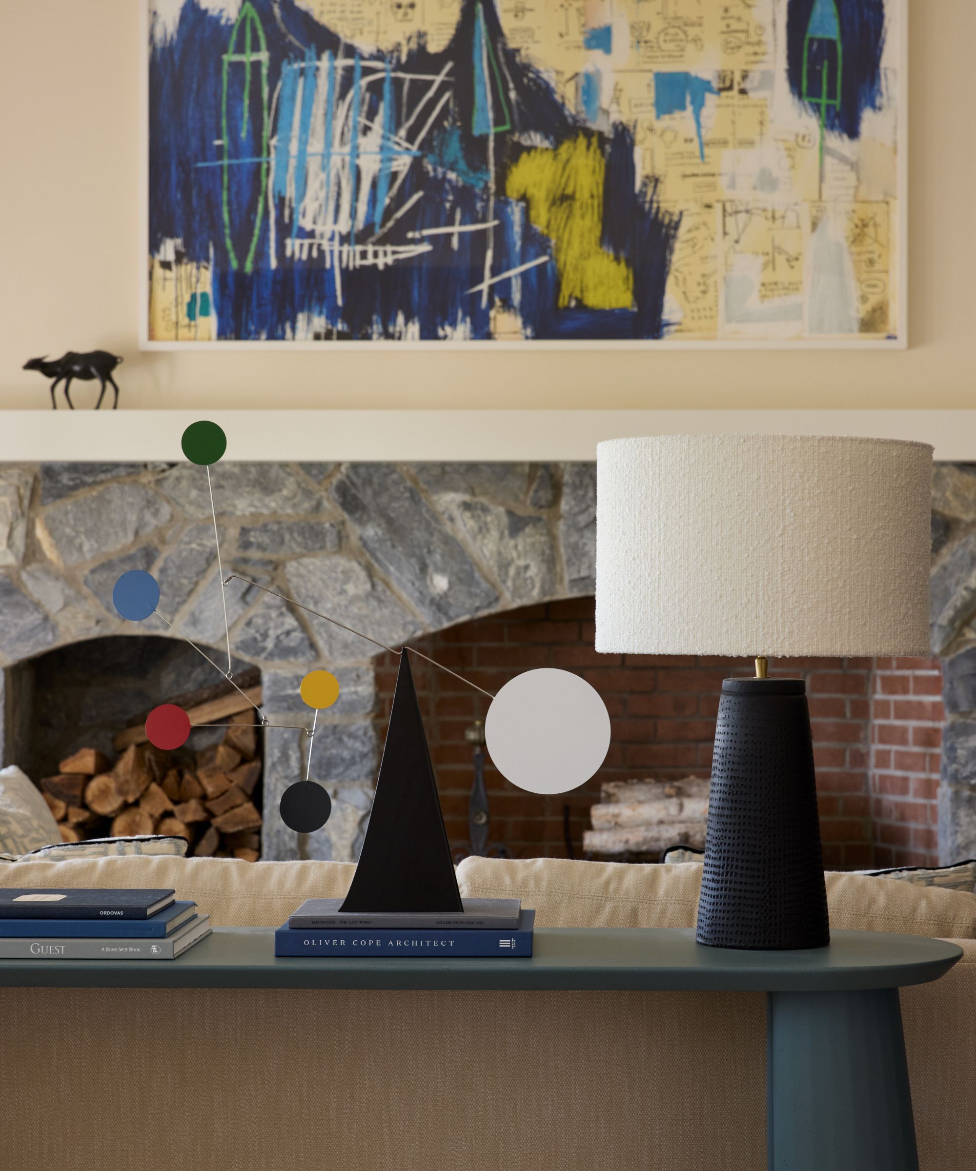 A cozy living room vignette featuring a stone fireplace with a modern abstract painting in blue, yellow, and black above the mantel, flanked by a black textured lamp and a colorful geometric sculpture on a light green console table