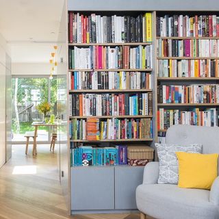 A grey chair in front of two large full height bookshelves full with books, showing a hallway leading to a kitchen area with table in front of large full height glass windows