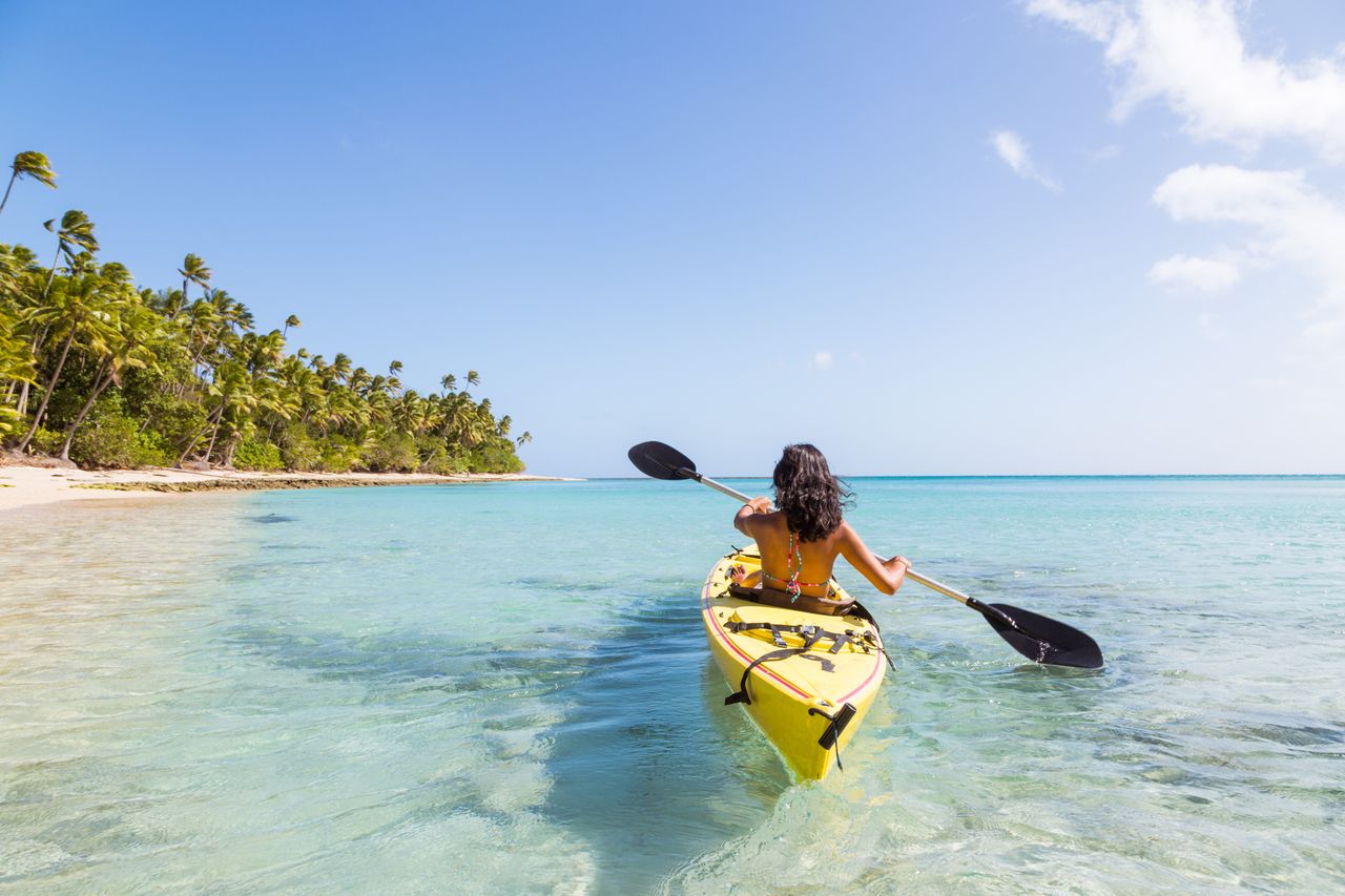 A woman kayaking away from the camera in shallow water off of a Fiji beach.