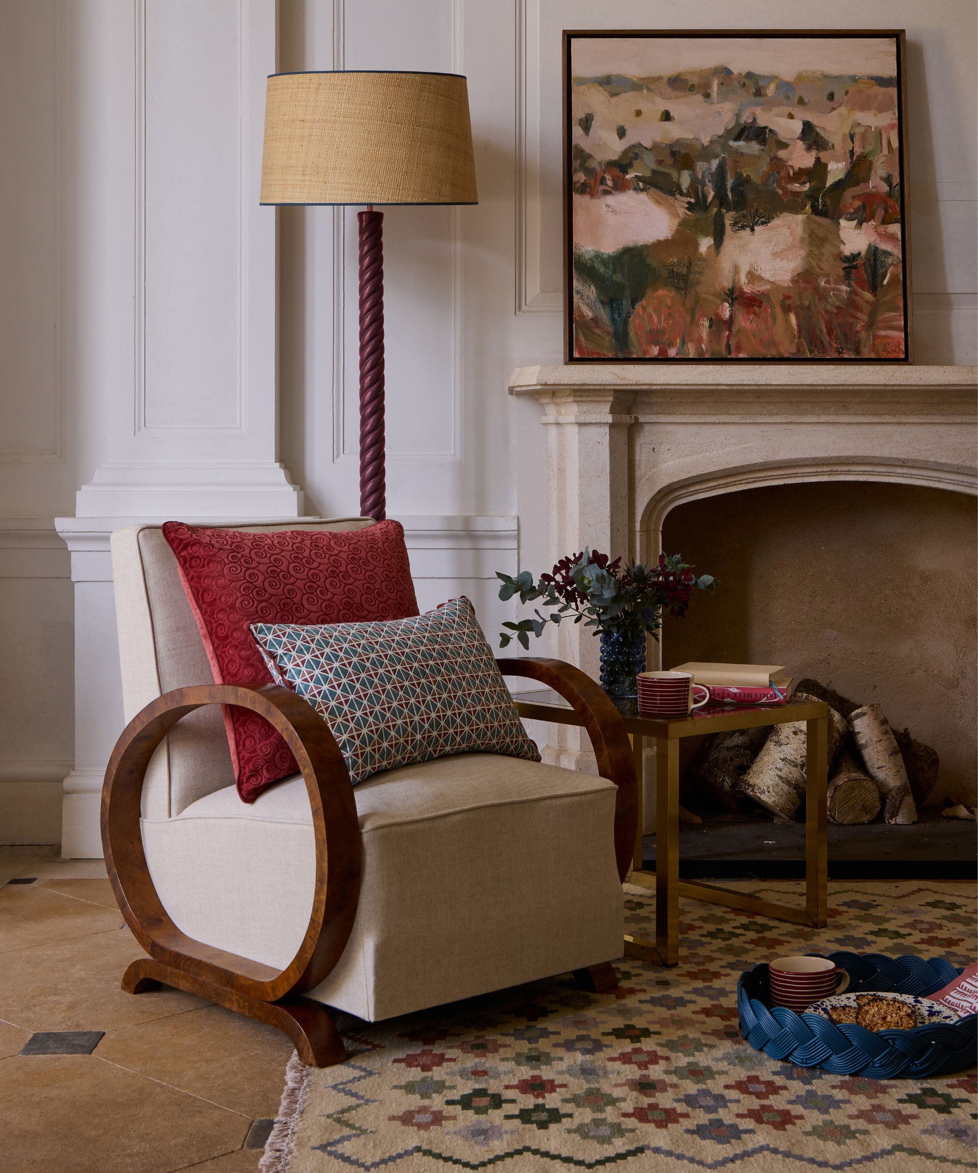 neutral living room in a period property with a large stone fireplace, with abstract art on the mantel. On the stone floor is a woven multi-color rug with a sculptural wooden armchair and a floor lamp with twisted details