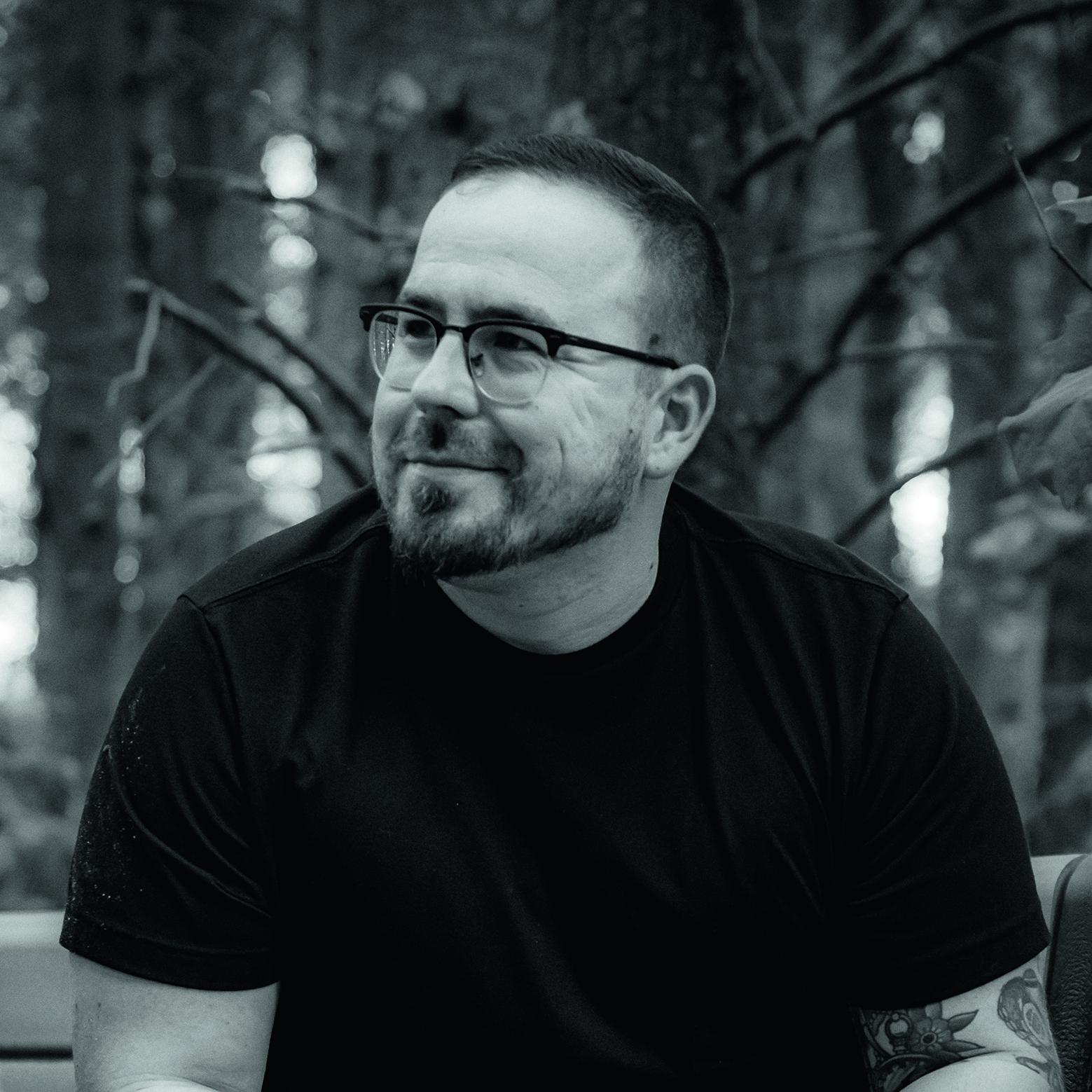 Black and white photo of a bearded man with glasses sitting outdoors, smiling softly. Forest background adds a calm, reflective atmosphere