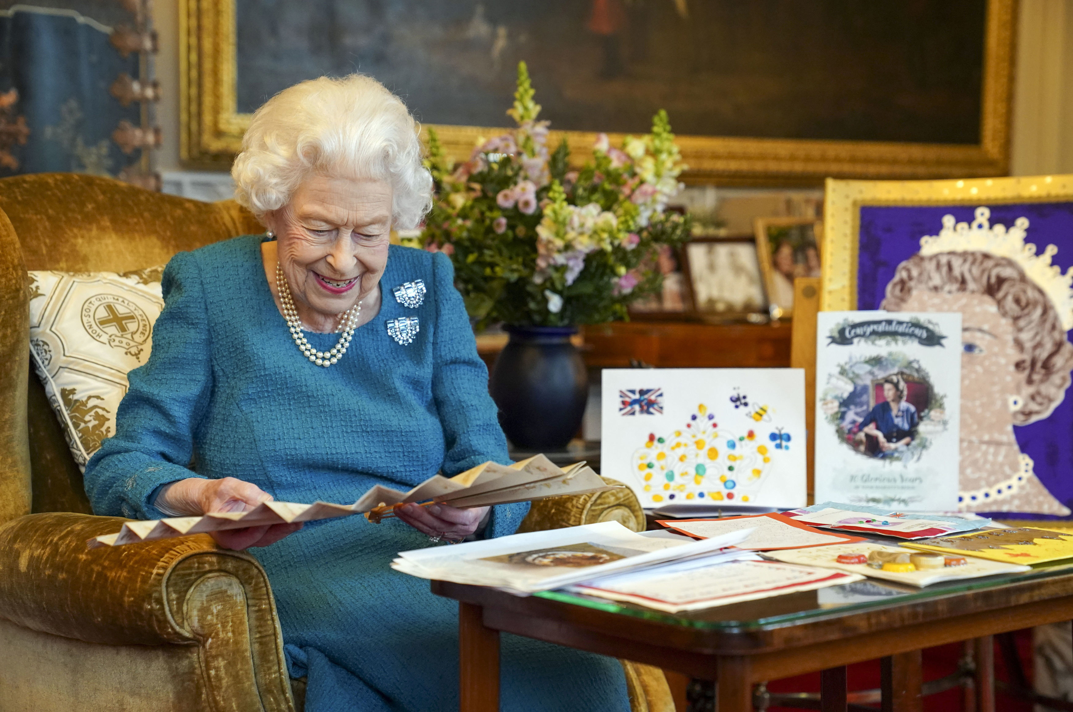 Queen Elizabeth wearing a blue dress smiling and looking at cards and a fan set on a table