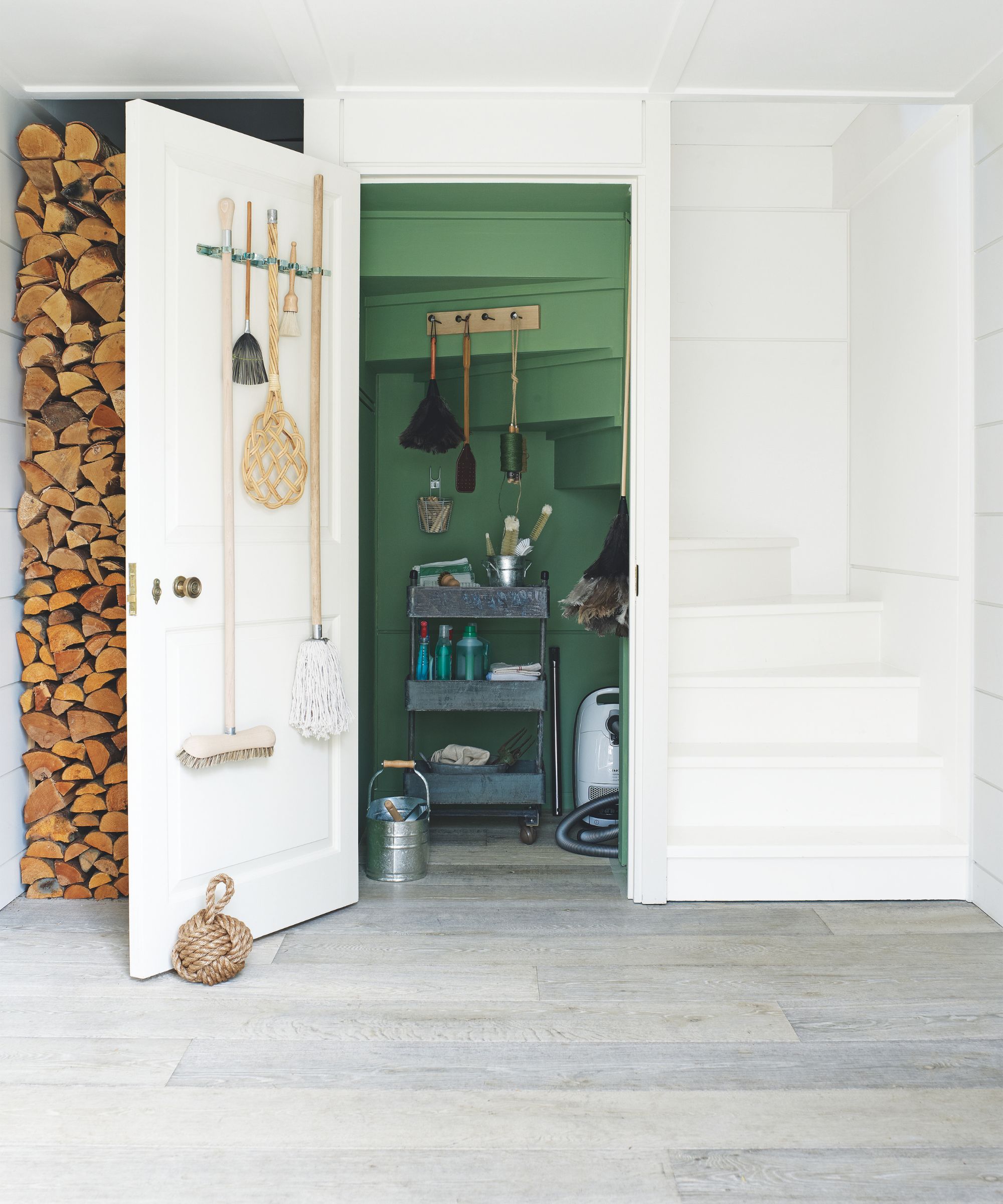 A green painted under-stairs cupboard filled with cleaning supplies and a rolling metal cart, with an open door, with various wooden cleaning tools hanging up. To the left of the open door is a tall logstore, and to the right is a white staircase. The floor is pale grey wood.