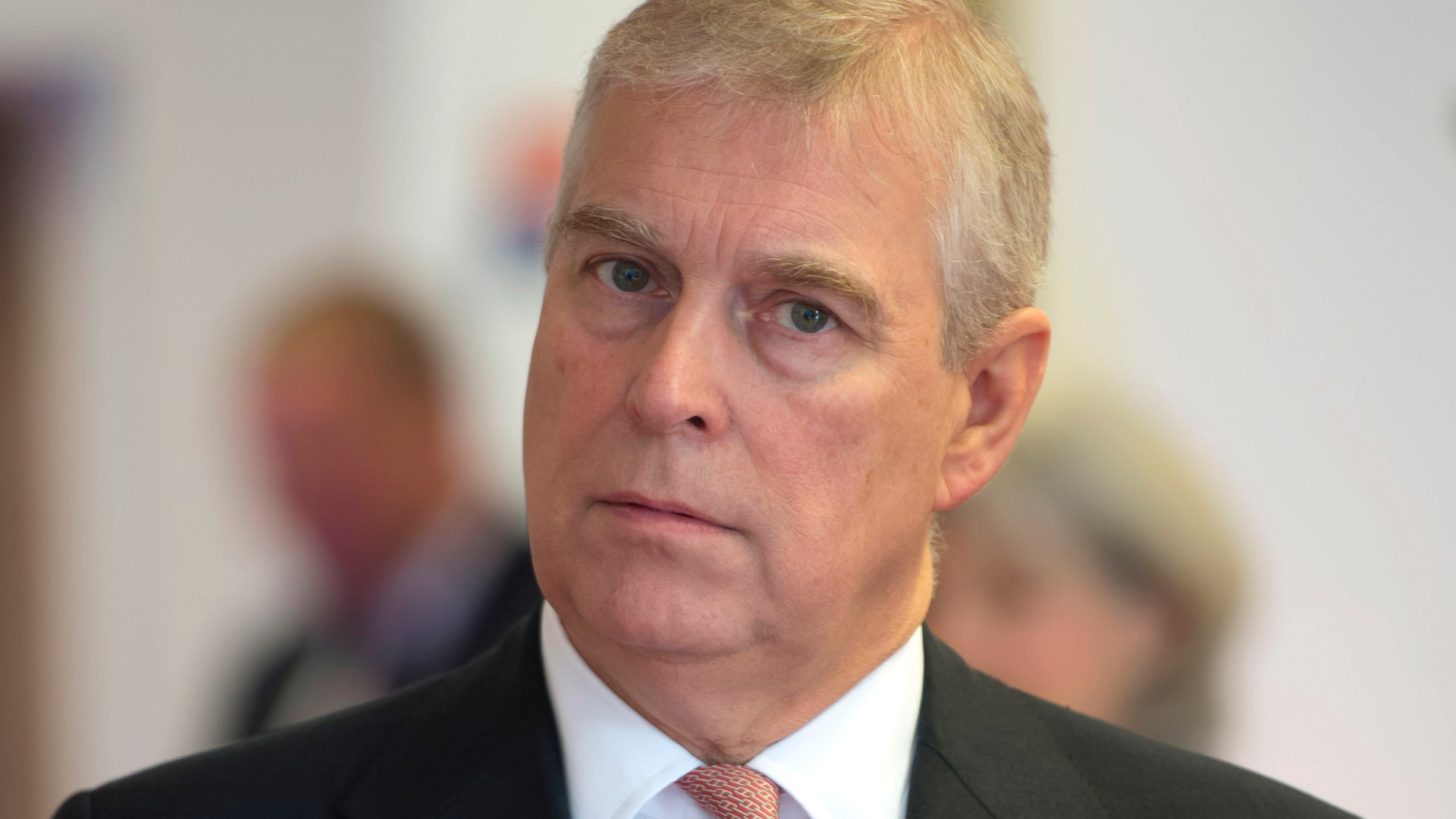The former Prince Andrew looks annoyed while wearing a black suit, a white shirt, and a red tie