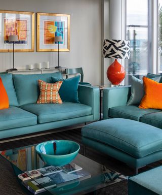 colors that go with teal, apartment living room with teal couches, footstool, glass coffee table, light grey walls, orange accents, artwork