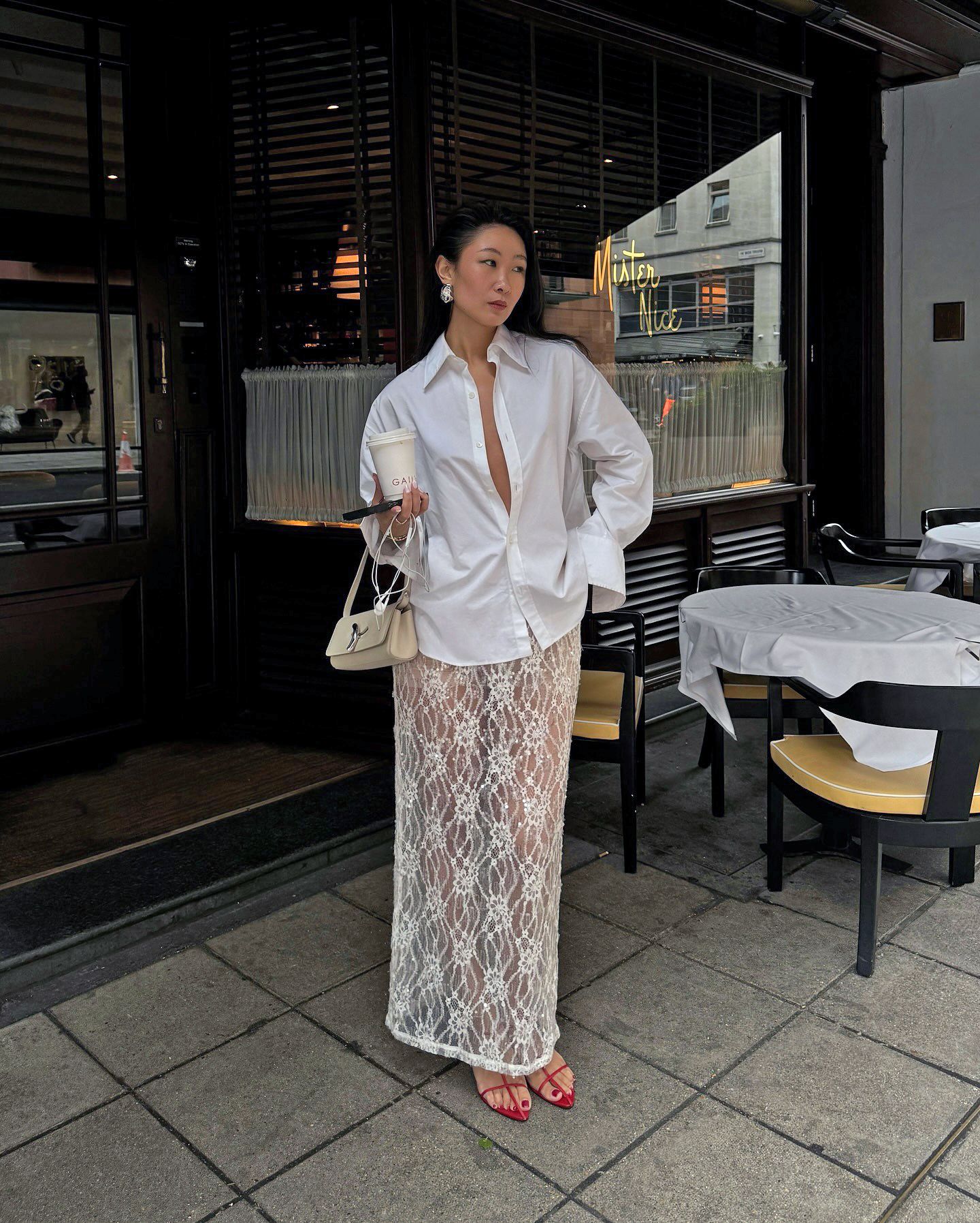 influencer Caroline Lin poses outside Mister Nice in London wearing an unbuttoned white button-down shirt with wide cuffs, small cream shoulder bag, white sheer lace maxi skirt and strappy red heeled sandals.