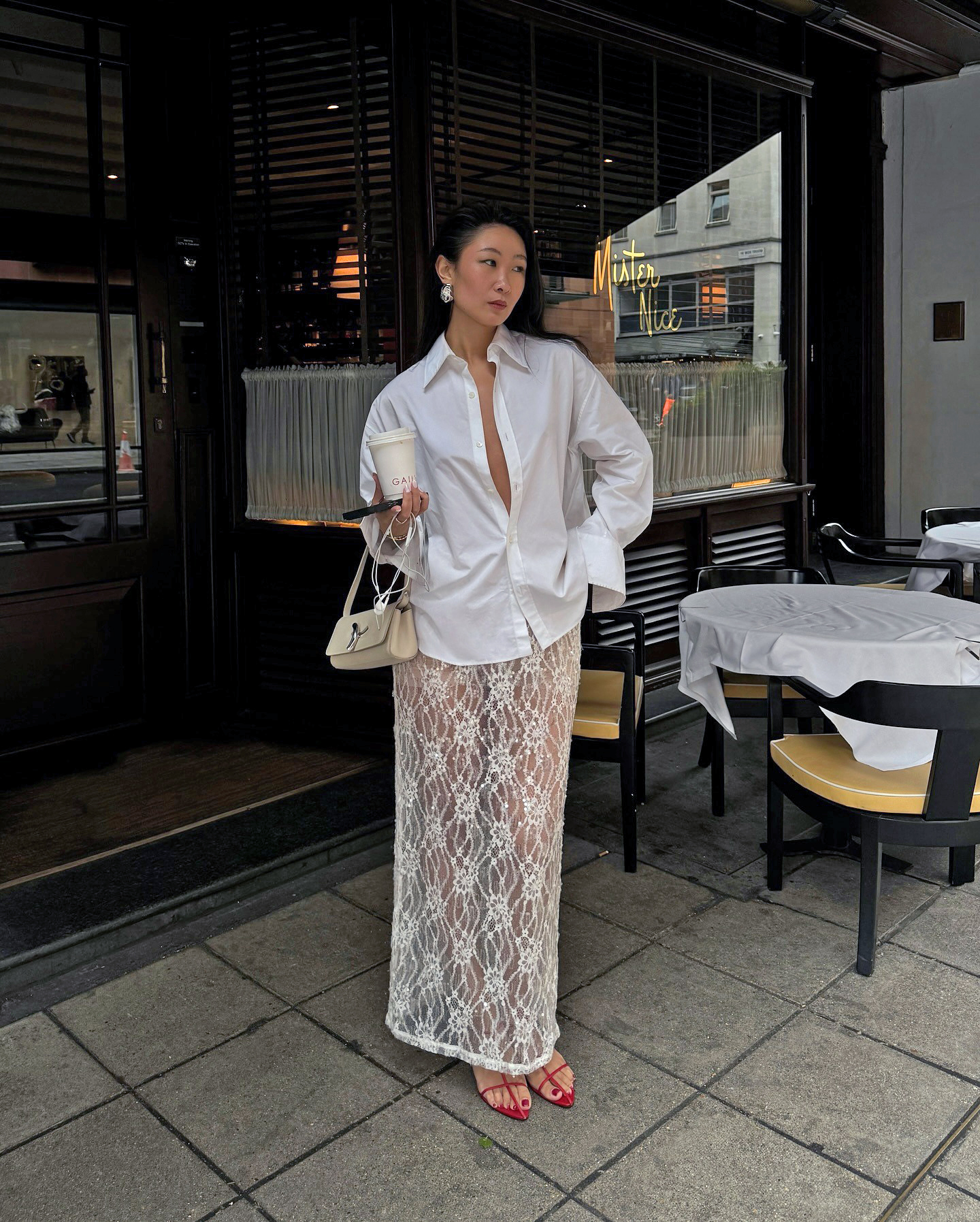 influencer Caroline Lin poses outside Mister Nice in London wearing an unbuttoned white button-down shirt with wide cuffs, small cream shoulder bag, white sheer lace maxi skirt and strappy red heeled sandals.