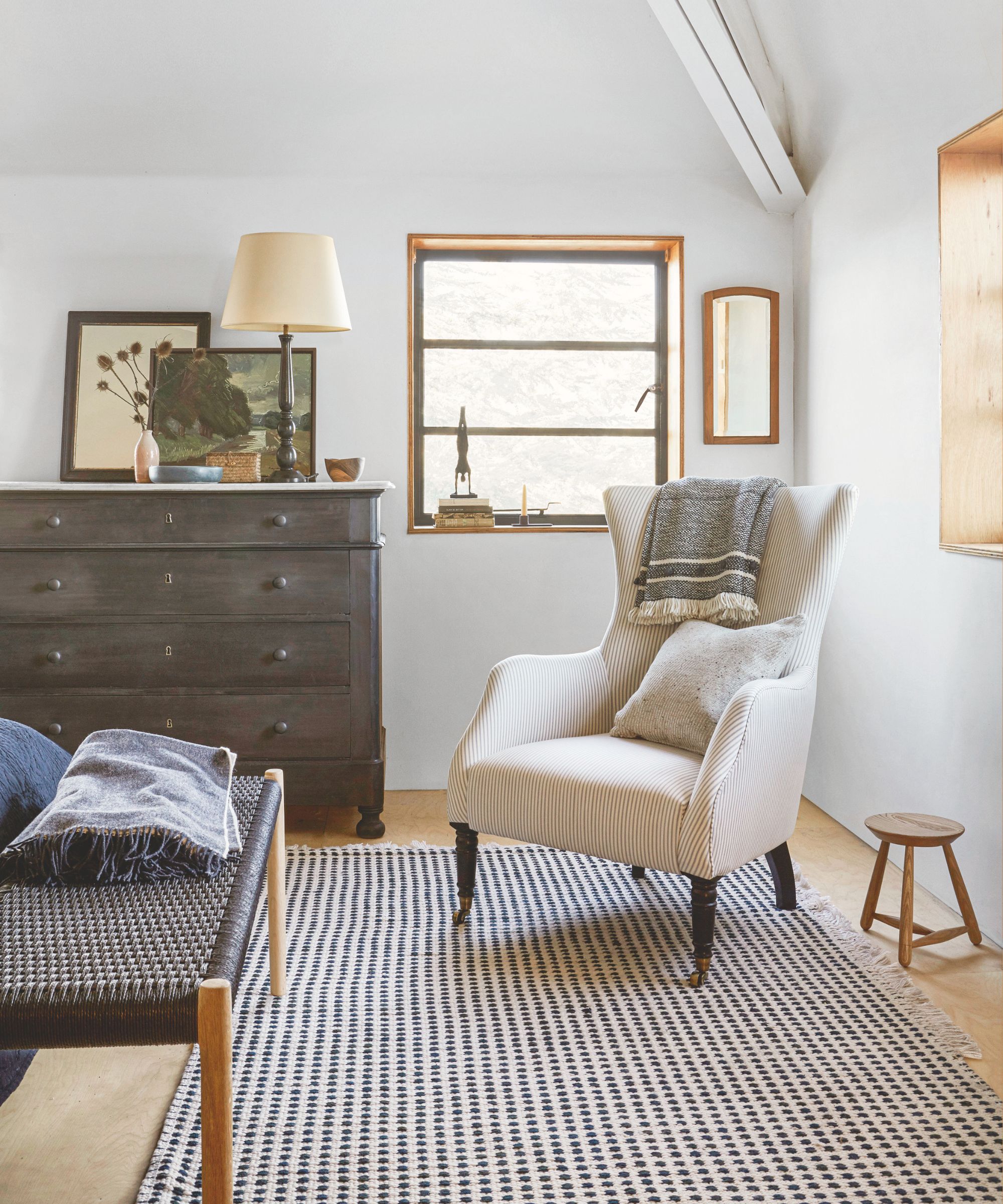 A neutral bedroom space with a black and white rug, white upholstered chair next to a small wooden stool, brown dresser with lamp and artworks and brown bench.
