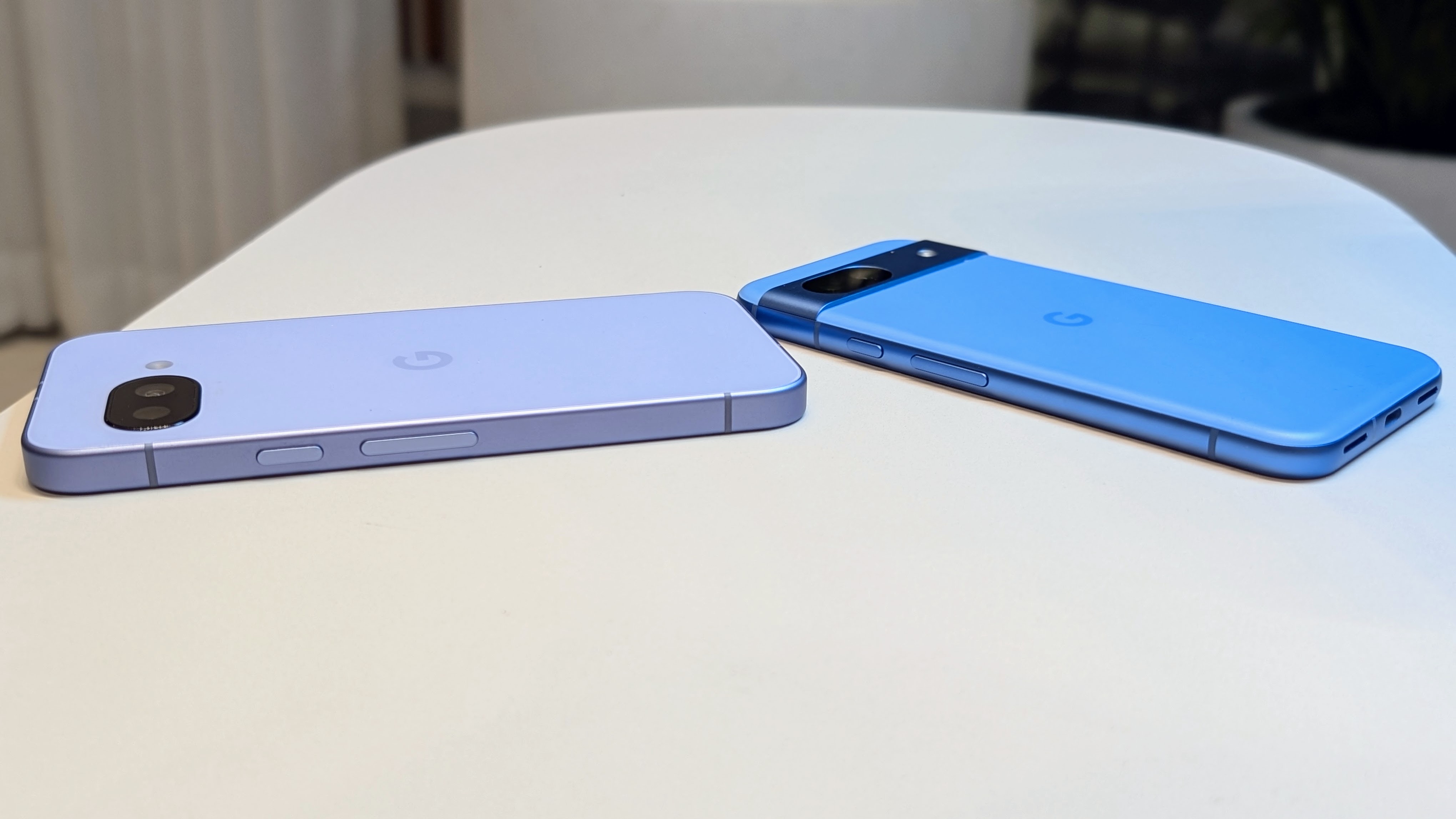 The Iris Google Pixel 9a (left) and Bay Google Pixel 8a (right) aside one another on a white table, the angle showing their relative difference in thickness.