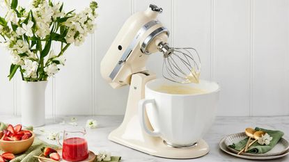 A cream Kitchenaid Artisan mixer with a white ceramic bowl flanked by a bouquet of white flowers, juice, and a set of plates