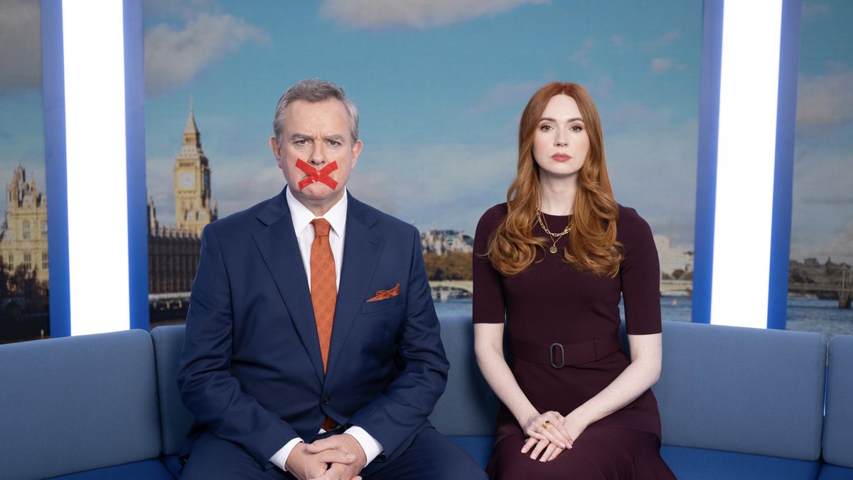 Hugh Bonneville as Douglas in a suit with a red cross taped across his mouth sits on a sofa next to Karen Gill as Madeline in a maroon dress against a London backdrop in Douglas is Cancelled