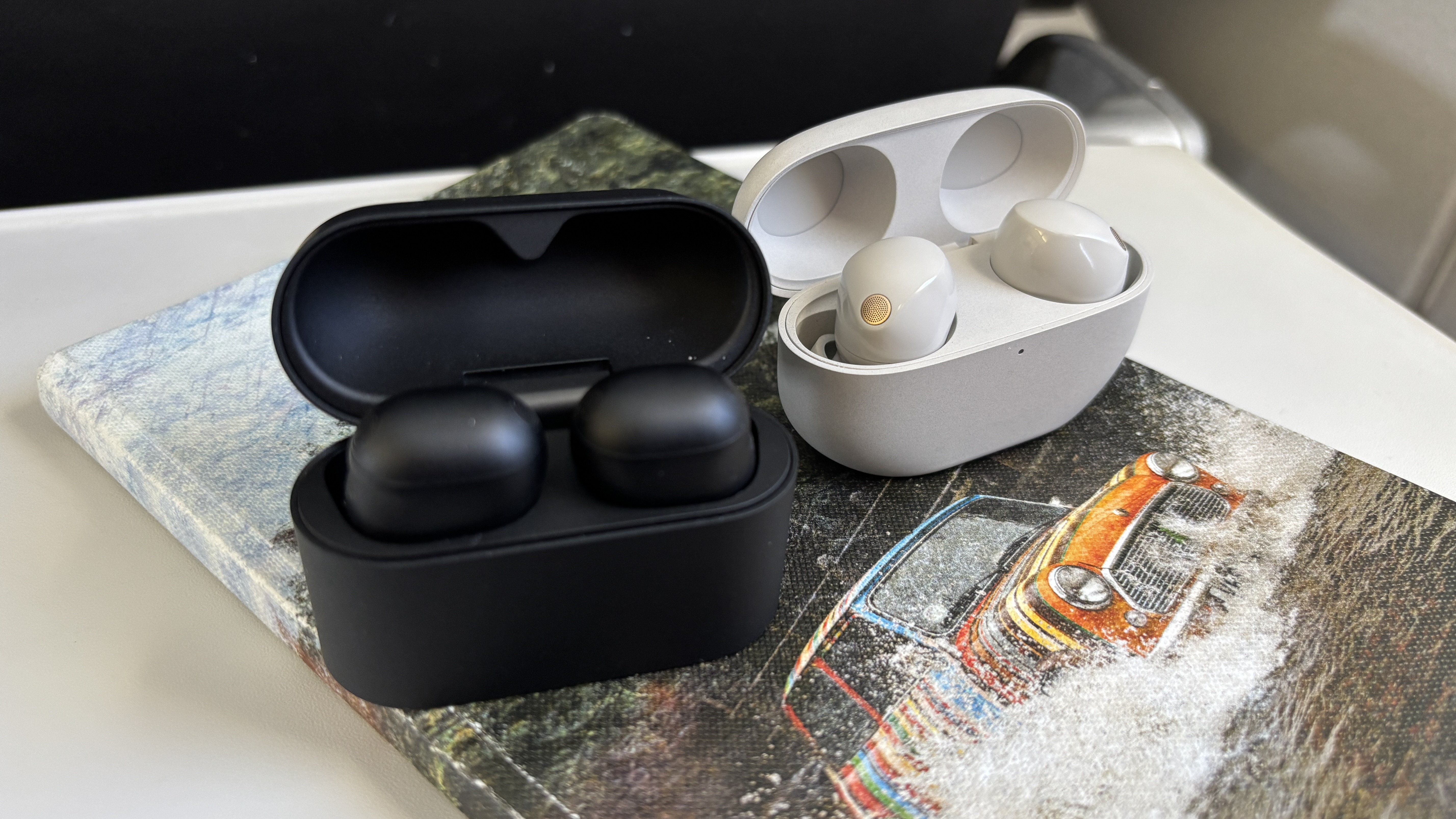 Sony WF-1000XM6 and XM5 wireless earbuds in their cases, with the lids open, placed on a notebook