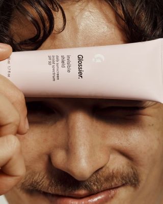 A man holds a pale pink moisturizer bottle in front of his forehead.