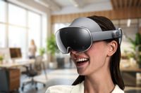 A smiling woman wears a grey mixed-reality headset with a glossy black front visor, captured in a bright, modern office setting.