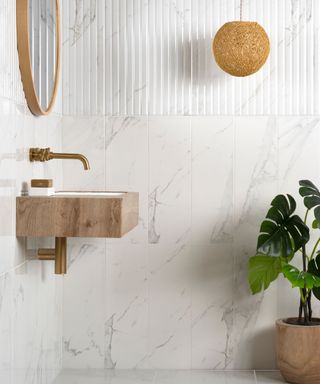 A minimalist bathroom featuring a wall-mounted wooden sink with a golden tap. The walls are clad in white marble-effect tiles with subtle grey veining, complemented by vertically grooved panelling for added texture. A round wooden mirror hangs above the sink.