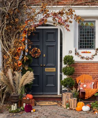 An autumnal porch with pumpkins and dried flowers around it and a house with a dark blue door and white walls